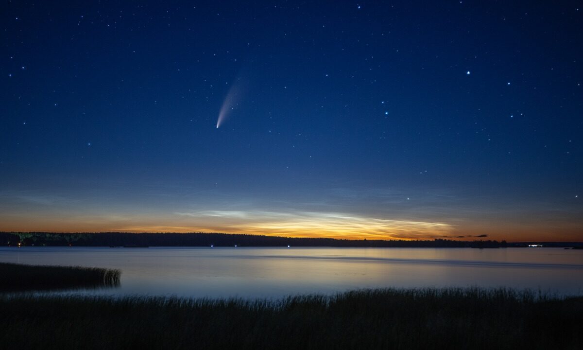 Kometa C/2020 F3 (NEOWISE) z jasnym warkoczem na granatowym niebie o zmierzchu, nad jeziorem i linią lasu na horyzoncie