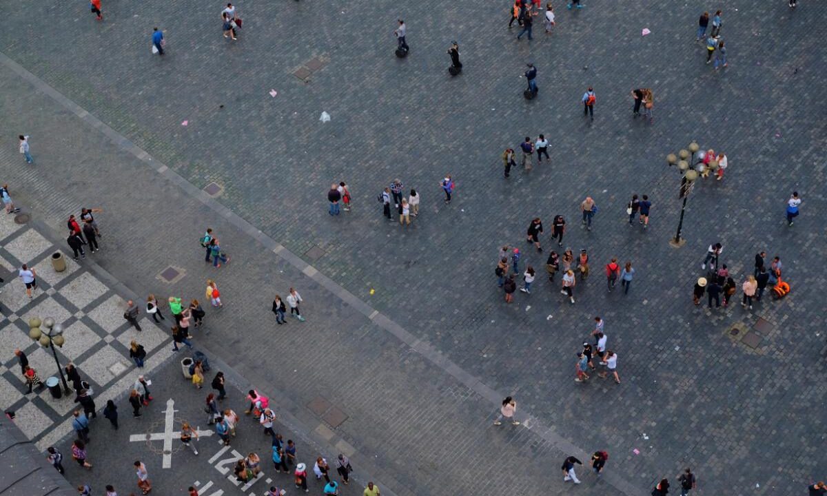 Widok z góry na szary plac miejski, po którym spacerują rozproszeni ludzie, przy krawędzi kostka brukowa w szachownicę