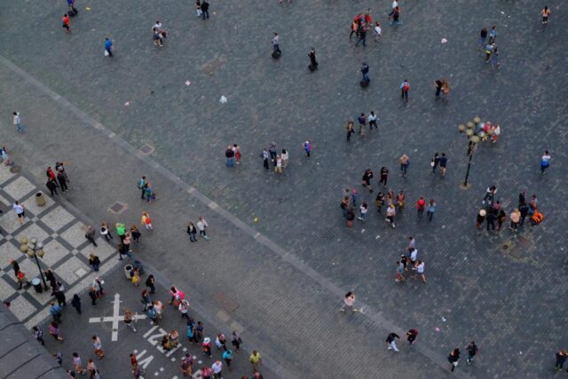 Widok z góry na szary plac miejski, po którym spacerują rozproszeni ludzie, przy krawędzi kostka brukowa w szachownicę